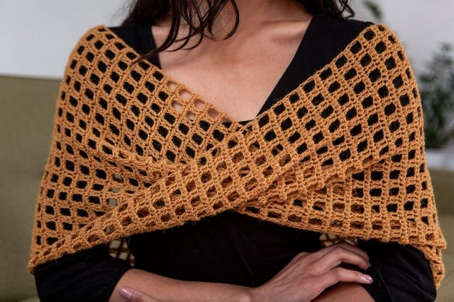 The Edith Wrap is a crochet shawl with an openwork square grid. This photo shows a dark yellow version of the shawl draped on the shoulders of a model.