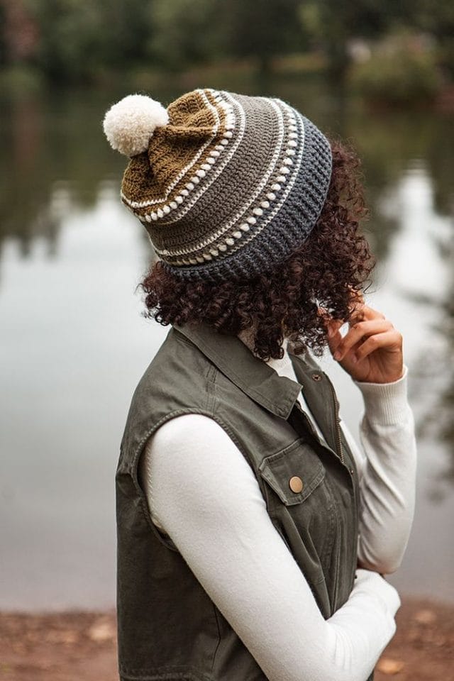 A model's head is turned to the side to display a crocheted hat in neutral colors with a white pom-pom. The pattern is Snow Mountain Hat, a free crochet pattern by crochet.com