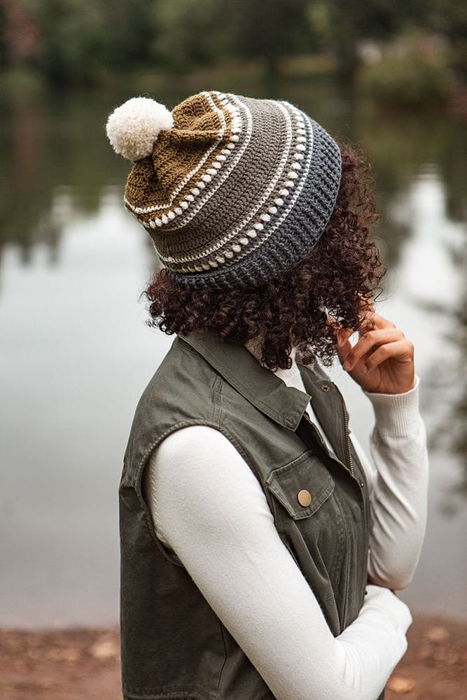 A model's head is turned to the side to display a crocheted hat in neutral colors with a white pom-pom. The pattern is Snow Mountain Hat, a free crochet pattern by crochet.com