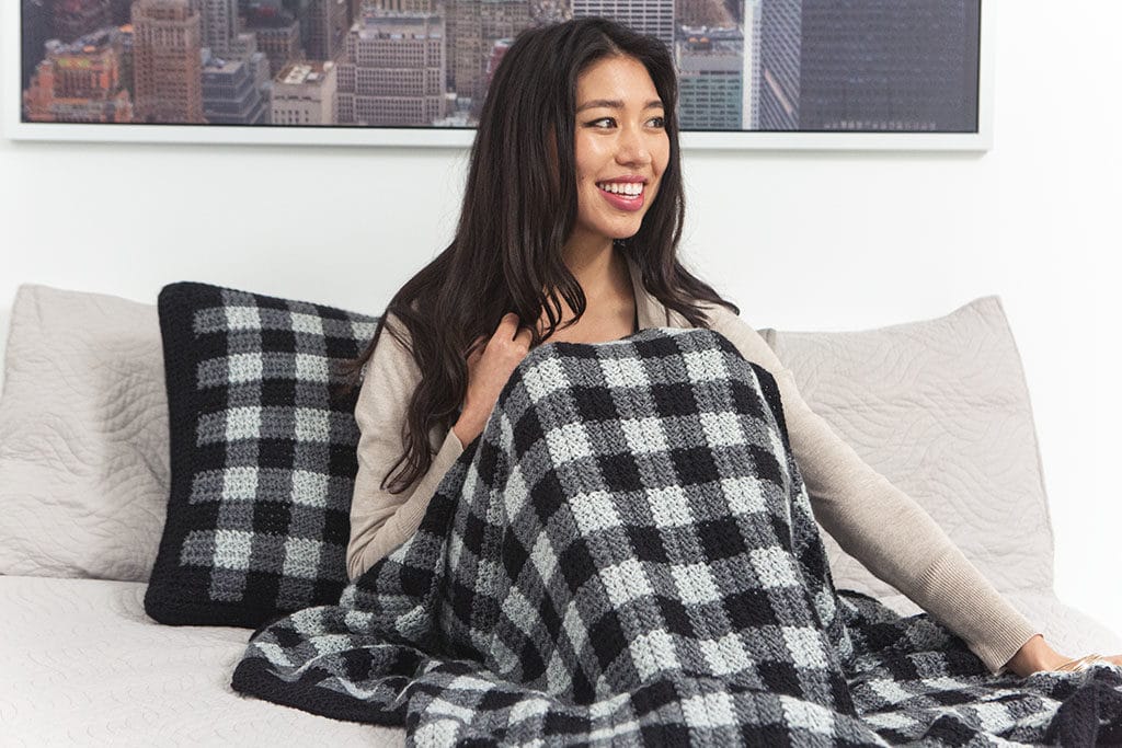 A woman sits on a bed under a plaid crocheted blanket