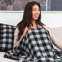 A woman sits on a bed under a plaid crocheted blanket