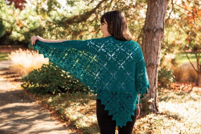 The Placid Shawl is a teal colored shawl made of square motifs, joined into a triangular shape. A model shows the shawl with its wingspan spread out.