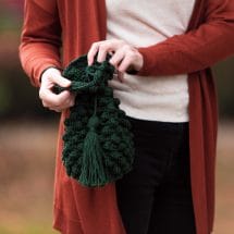 A hand holding a crocheted green bag with a drawstring top, big tassel, and a textured bobble-stitched body