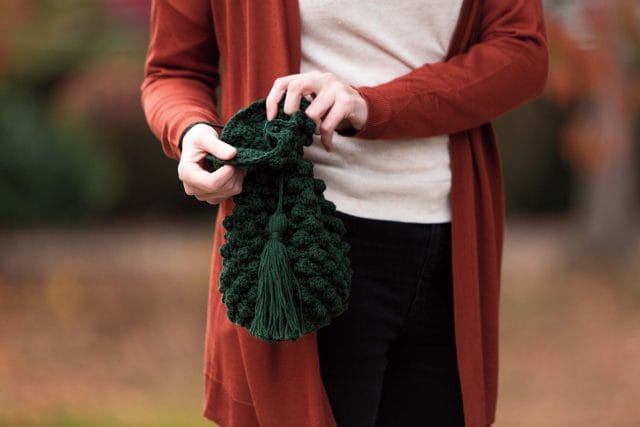 A hand holding a crocheted green bag with a drawstring top, big tassel, and a textured bobble-stitched body