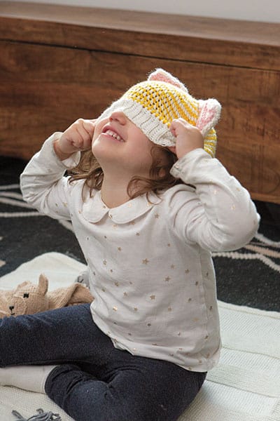 A little girl pulls a crochet hat with cat ears down over her face.