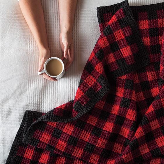 A red and black plaid crocheted blanket  draped on a bed, next to hands holding a coffee cup