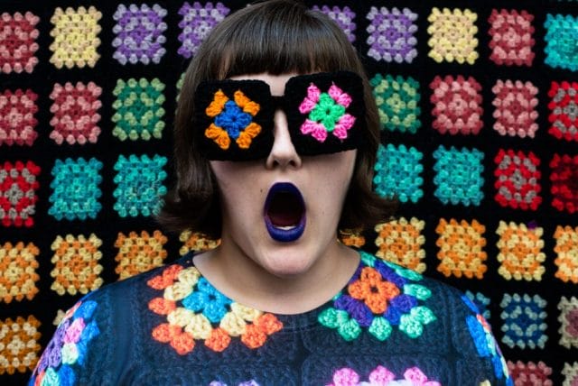 Ashley stands in front of a granny square afghan backdrop, wearing glasses with granny squares on the lenses, and a granny square printed top.
