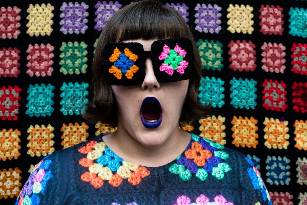 Ashley stands in front of a granny square afghan backdrop, wearing glasses with granny squares on the lenses, and a granny square printed top.