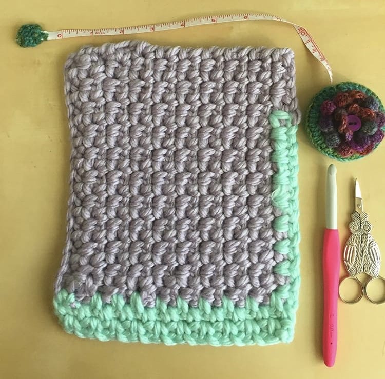 A rectangle of crochet in gray and mint next to a cute tape measure, owl-themed scissors, and crochet hook