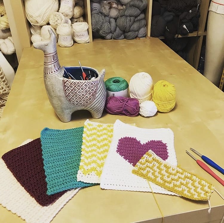 Six crochet rectangles in varying colors and patterns, one with a purple heart, in front of a llama-shaped planter filled with crochet hooks.