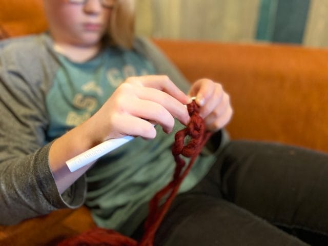 An image of a child's hands holding a crochet hook and bulky yarn. One tip for crocheting with kids is to use smooth yarn and a large hook.