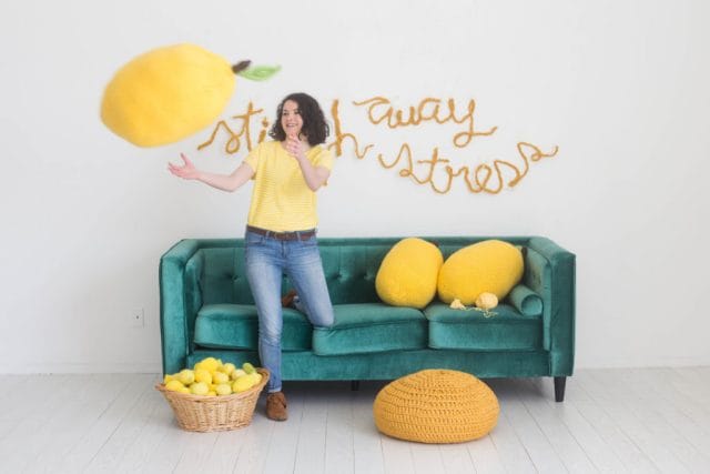 A woman tossing a crochet pillow in the shape of a lemon in front of a green couch covered in knit and crochet lemon pillows and the words stitch away stress on the wall.