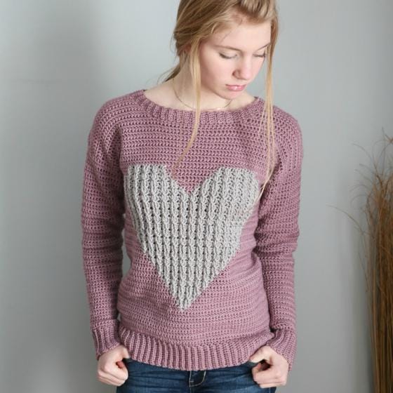 A girl models a pink crocheted sweater with a big textural gray heart on it.