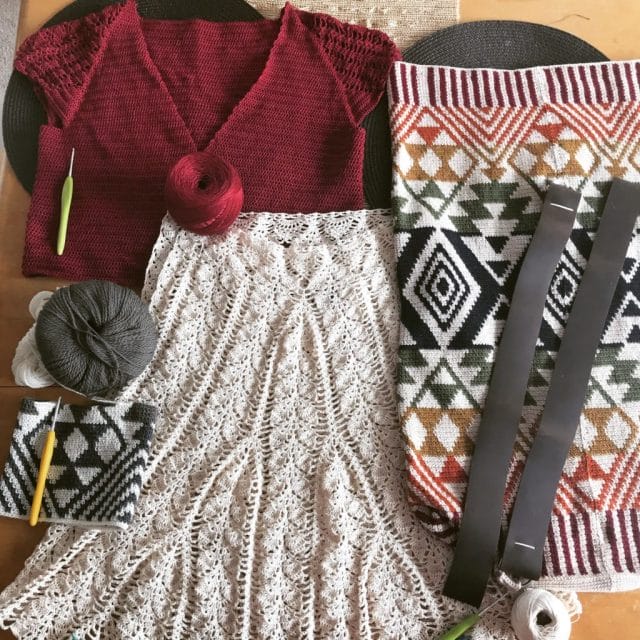 A top-down view of a table covered in crochet projects and yarn: A red top made of Curio crochet thread, a cream-colored skirt made in Curio crochet thread, a tapestry crochet backpack.