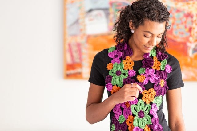 a model wears a scarf made of crocheted flowers in purples, oranges, and greens
