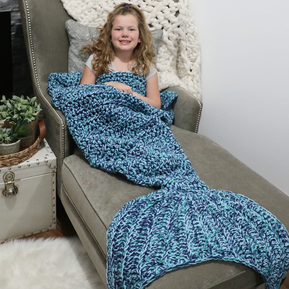 A little girl lounges on a couch, wearing a large mermaid tail blanket.
