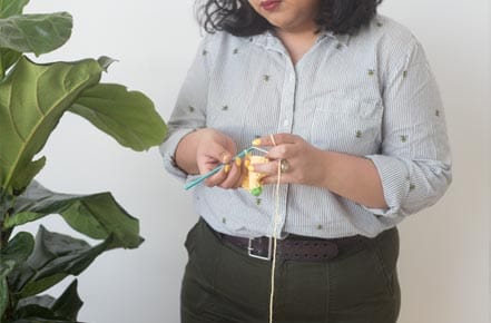 A woman crochets with yellow yarn next to a leafy plant.