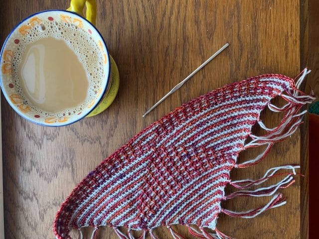 A top-down view of a tabletop featuring a cup of coffee, a crochet hook, ,and a triangular swatch of a crochet project.
