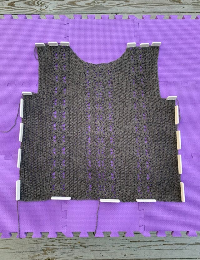 Half a gray crocheted sweater blocking on purple blocking mats, set on a wooden deck.