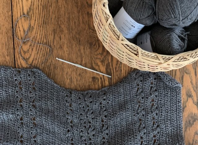 A wooden background with a blonde basket full of gray yarn on the right. The top half of a crocheted sweater in gray, accompanied by a metal crochet hook.