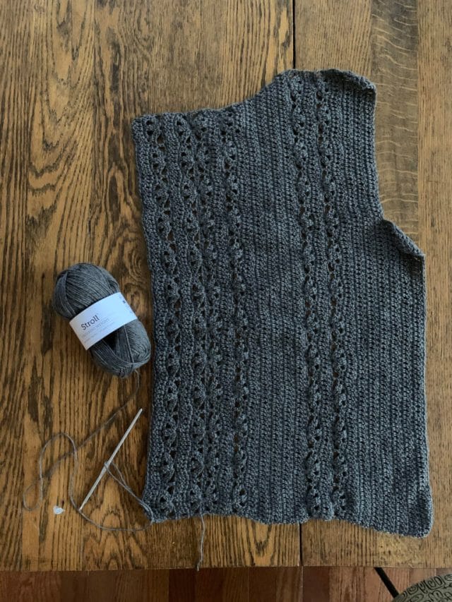 A wooden background with a half of a crocheted sweater in gray, accompanied by a ball of gray Stroll yarn and a metal crochet hook.