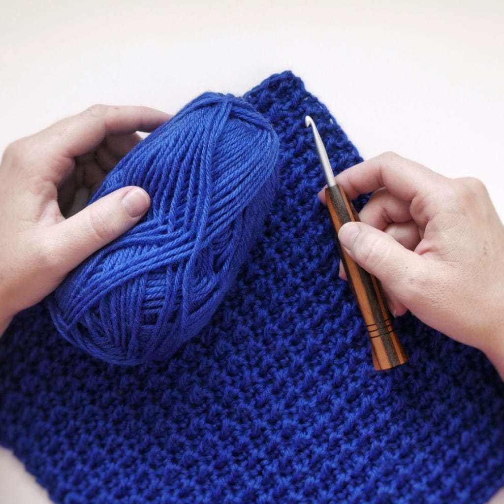 Hands holding a ball of blue yarn and a wooden handled crochet hook over a blue crocheted swatch with a bumpy texture.