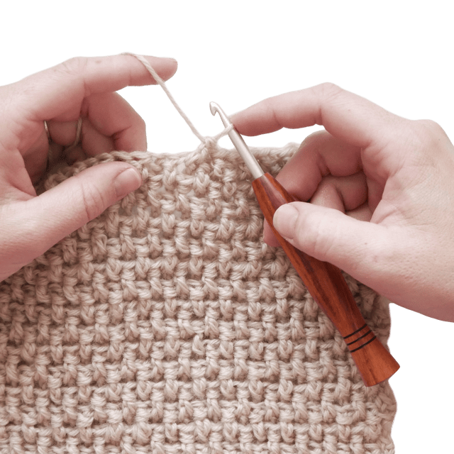 An image of hands crocheting a tan crochet swatch with a basketweave texture, with a wooden-handled crochet hook.