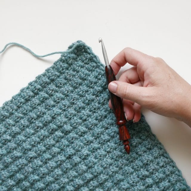 A hand holding a wooden hook over a greenish-blue textured crochet swatch on a white background. 