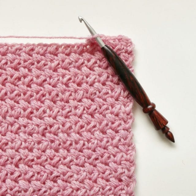 A pink textured crochet swatch on a white background. A wooden crochet hook is hooked into the top of the swatch.