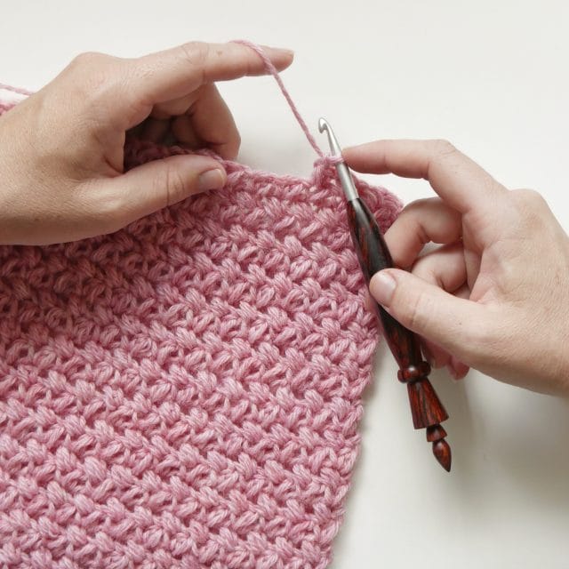 Hands crocheting with a wooden crochet hook into a pink textured crochet swatch on a white background. 