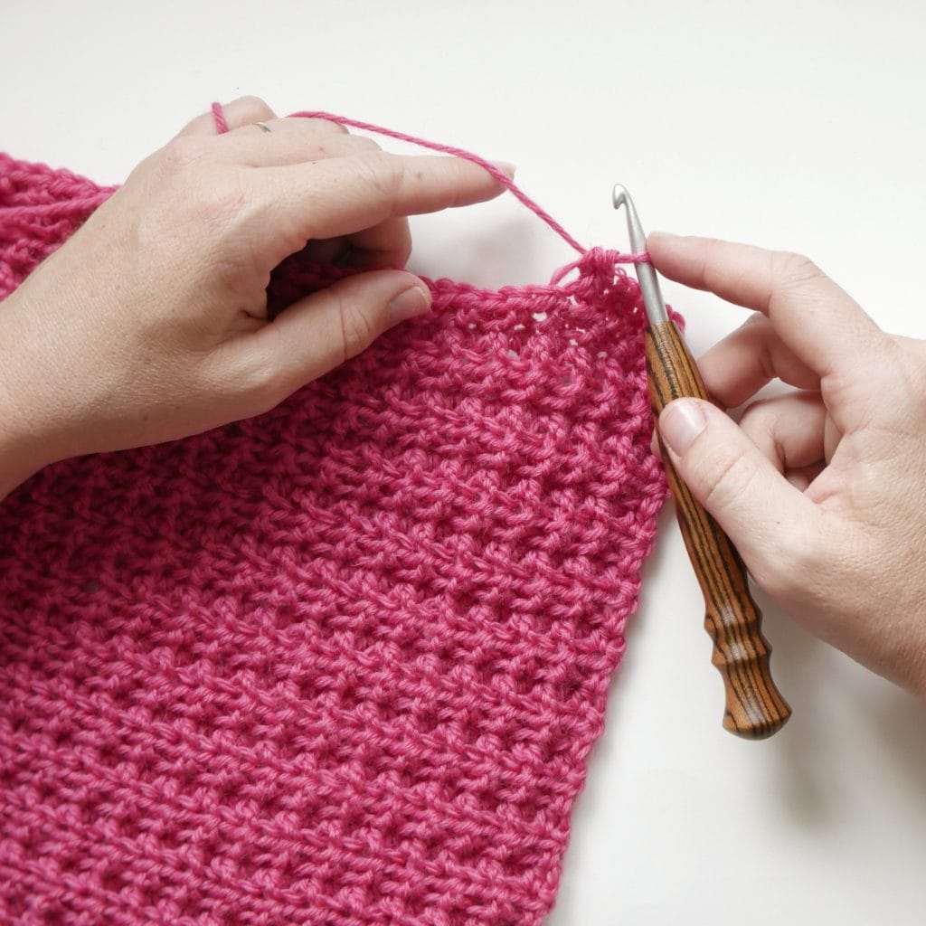 Hands holding a wooden crochet hook crocheting a bright pink crochet swatch with a waffle-ish texture on a white background. 