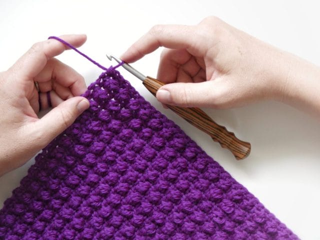 Hands crocheting with a striped wooden crochet hook, on a white background: A purple crochet swatch featuring a textured stitch (the even berry stitch).
