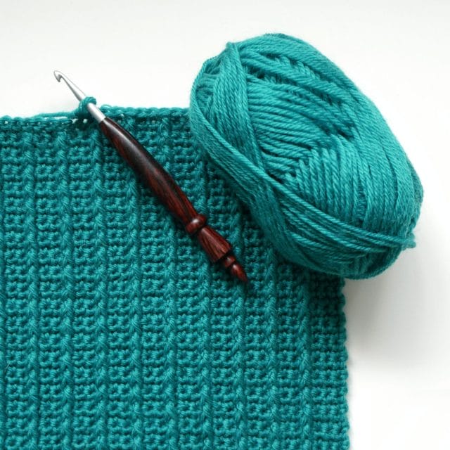 An image of a turquoise crocheted swatch with vertical lines of front post double crochet spanning up and down the swatch. A wooden crochet hook lays across the swatch, and a ball of yarn sits on the corner.