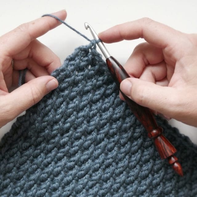 Hands crocheting with a wooden hook, along the corner of a blue-gray crochet swatch with nubby texture.