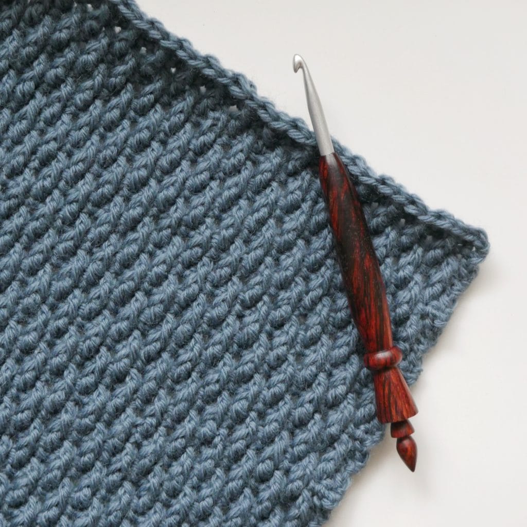 The corner of a blue-gray crochet swatch with nubby texture on a white background, with a wooden hook laying across the edge of the swatch.