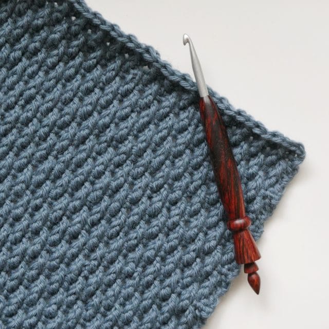 The corner of a blue-gray crochet swatch with nubby texture on a white background, with a wooden hook laying across the edge of the swatch.