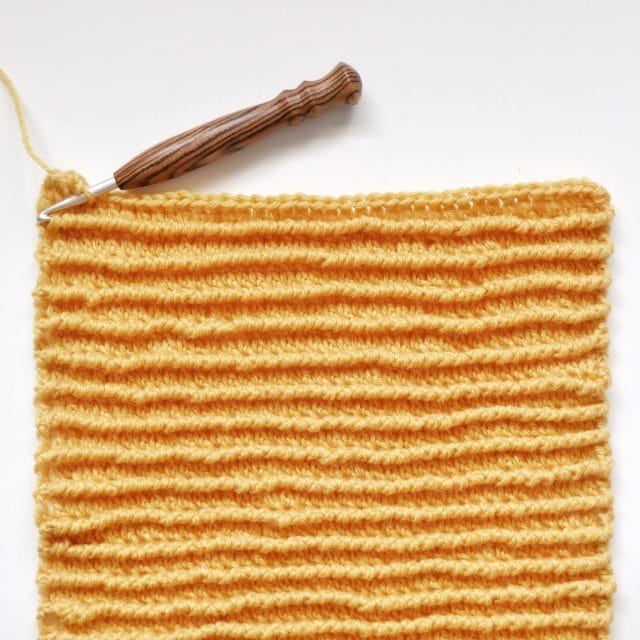 A striped wooden hook is set on top of  a bright yellow crochet swatch with a horizontal ribbed texture on a white background. 