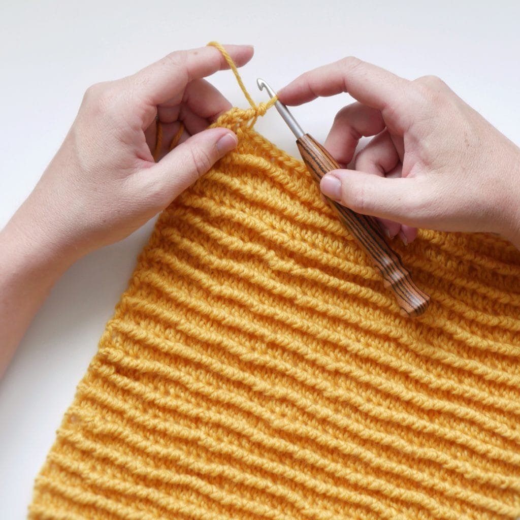 Hands crocheting with a striped wooden hook onto a bright yellow crochet swatch with a horizontal ribbed texture on a white background. 