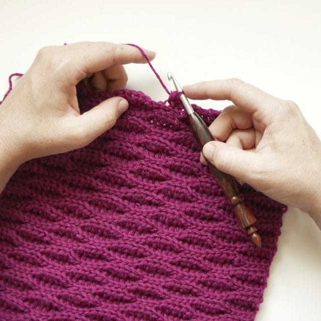 Hands crocheting with a wood-handled crochet hook onto a bright pink/fuchsia crochet swatch with an intriguing wave-like texture.
