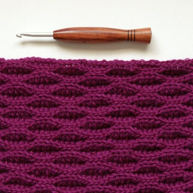 A wood-handled crochet hook sits above a bright pink/fuchsia crochet swatch with an intriguing wave-like texture on a white background.