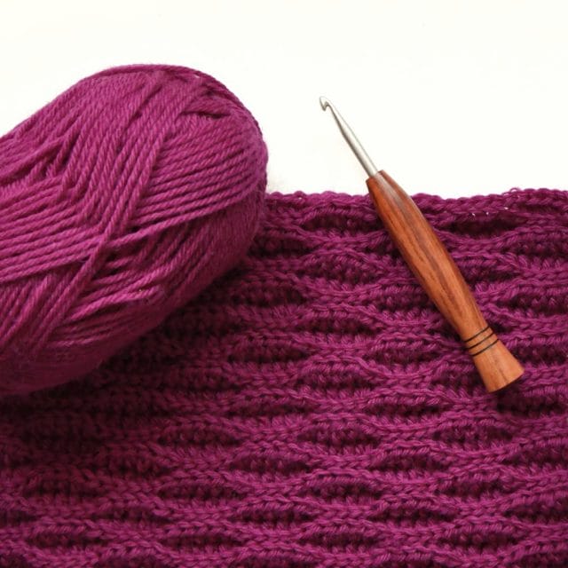 A ball of bright pink/fuchsia ball of yarn and a wood-handled crochet hook sits atop a bright pink/fuchsia crochet swatch with an intriguing wave-like texture on a white background.