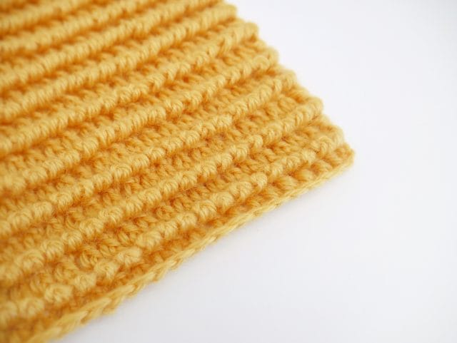 The corner of a bright yellow crochet swatch with a horizontal ribbed texture on a white background.