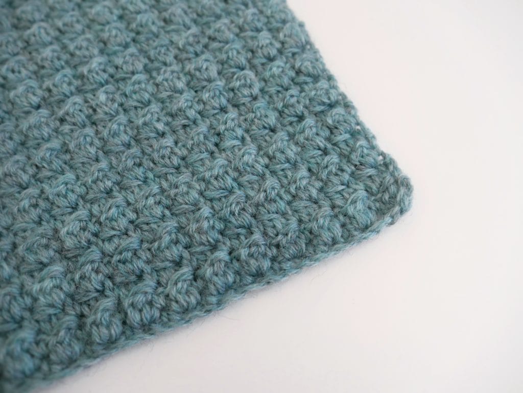 The corner of a greenish-blue textured crochet swatch on a white background. 