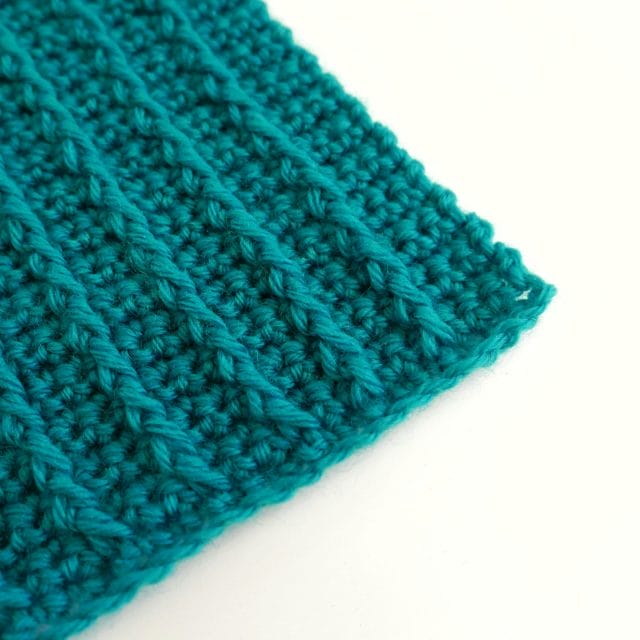 An image of a turquoise crocheted swatch (sitting diagonal across the image) with vertical lines of front post double crochet spanning up and down the swatch.
