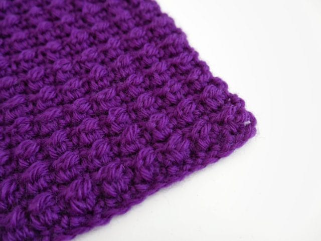 On a white background: the corner of a purple crochet swatch featuring a textured stitch (the even berry stitch).
