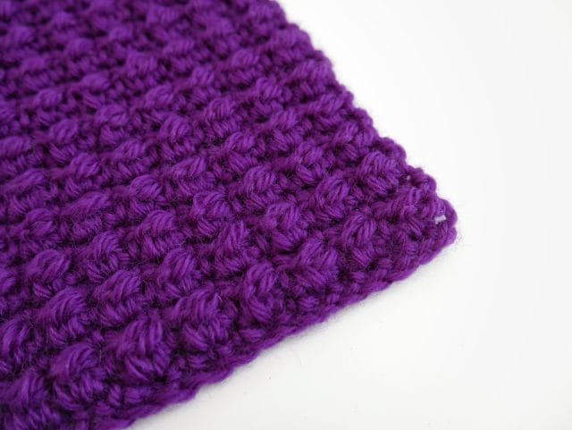 On a white background: the corner of a purple crochet swatch featuring a textured stitch (the even berry stitch).