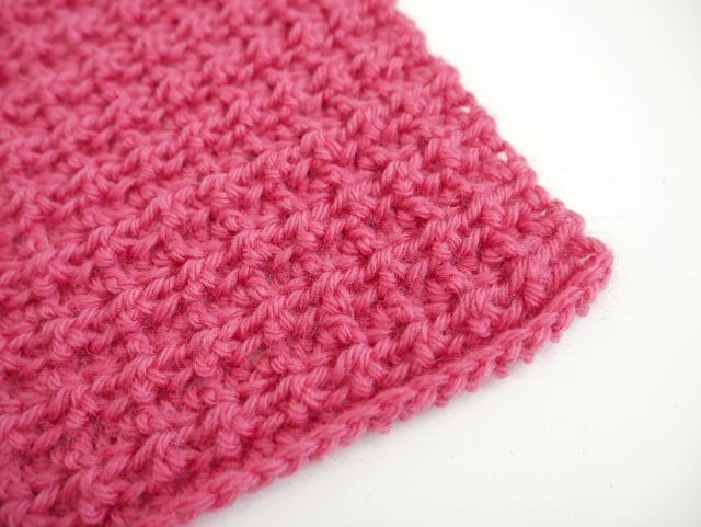 The corner of a bright pink crochet swatch with a waffle-ish texture on a white background. 