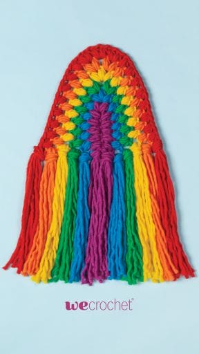 On a sky-blue background, a crocheted rainbow wall hanging, reminiscent of macrame, in rainbow colored puff stitches: red, orange, yellow, green, blue, purple. 