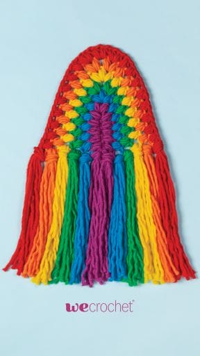 On a sky-blue background, a crocheted rainbow wall hanging, reminiscent of macrame, in rainbow colored puff stitches: red, orange, yellow, green, blue, purple.