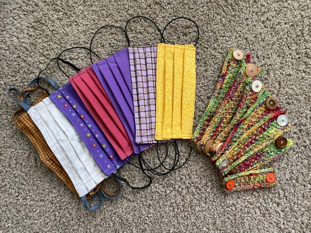 A top-down view of a pile of handmade fabric face masks with elastic straps, next to a pile of crocheted ear savers (4" x 1" rectangles with buttons at each end)